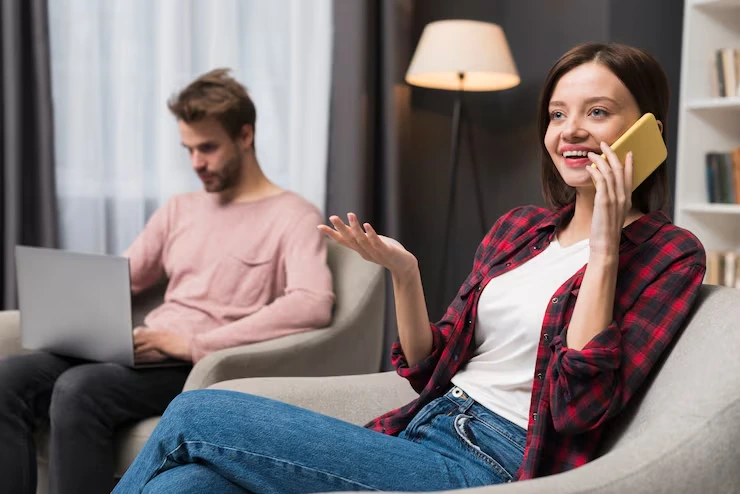 A Smiling Woman Receiving A B2C Cold Call At Home While A Man Works On A Laptop In The Background